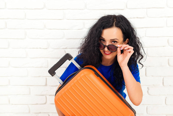 Happy woman in sunglasses holding travel suitcase. Emotional girl before trip isolated on white brick wall. Lifestyle, travel and vacation concept. Young woman holds orange suitcase. Copy space - Photo, Image