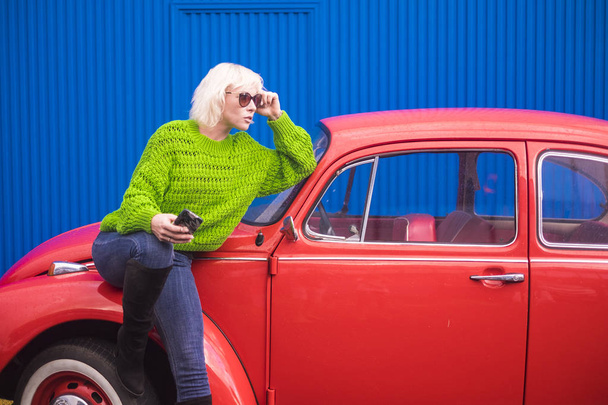 Moda y atractiva mujer blanca cabello joven con teléfono móvil llamando a amigos o revisando Internet con coche vintage rojo en segundo plano. - Foto, imagen