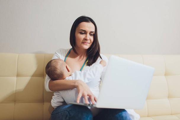 Beautiful young mother working with laptop computer and breastfeeding her newborn baby at home. Mom - business woman. - Фото, изображение