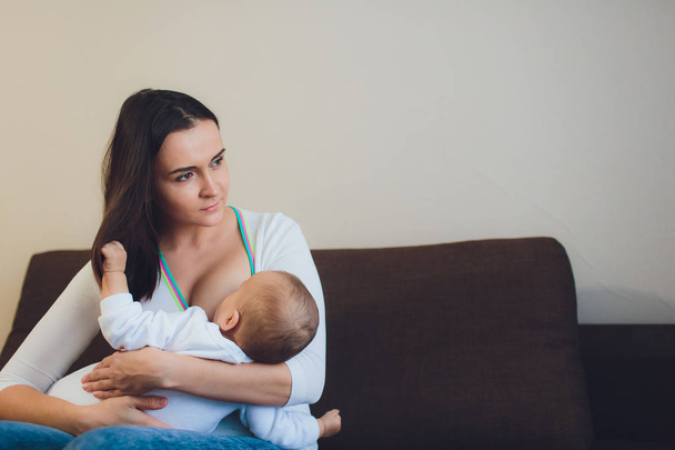 A Mother breastfeeding her little baby boy in her arms. brown sofa, home, white top - Photo, image