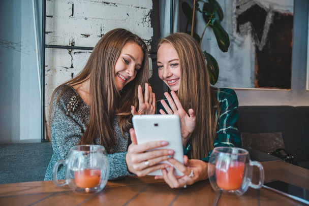 Две юные красавицы разговаривают в видеочате с помощью планшета. сидя в кафе, попивая чай
 - Фото, изображение