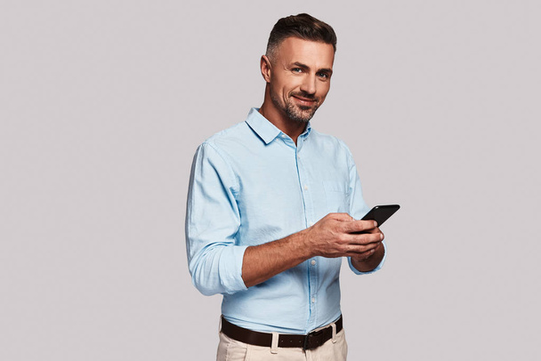 handsome man holding smart phone and smiling while standing against grey background - Фото, изображение
