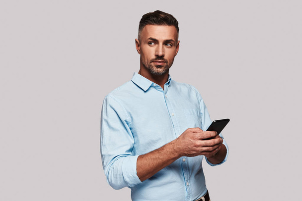 man in smart casual wear using his smart phone and smiling while standing against grey background - Zdjęcie, obraz