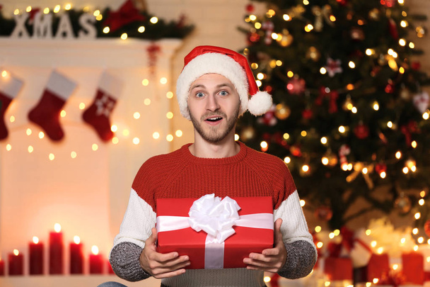 Young man in santa hat with gift box near christmas tree at home - Фото, изображение