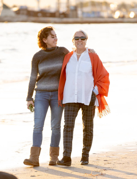Portrait of happy senior mother and adult daughter spending time together holding hands laughing and walking on beach at sunset light in Happy family moments Generations Retirement and People concept. - Photo, Image