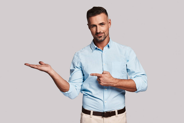 handsome young man pointing hands for copy space and looking at camera while standing against grey background - Foto, immagini