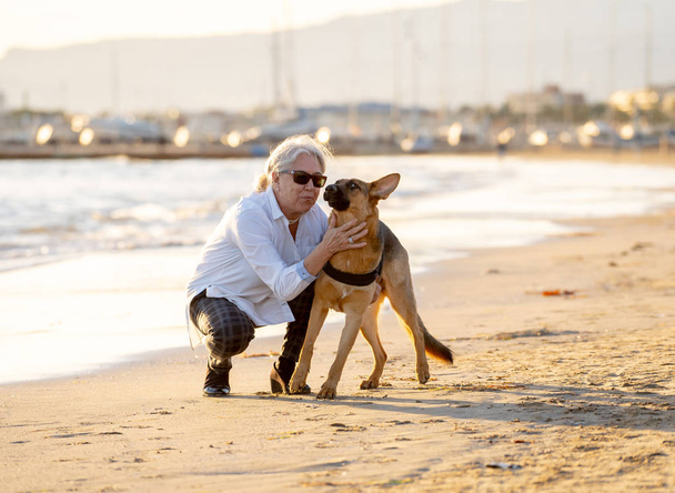 Beautiful widow older woman playing with her german shepard dog enjoying its companion and love on beach at sunset in Benefits of animals Keeping active Retirement lifestyle and Dog friendly tourism. - Foto, Bild