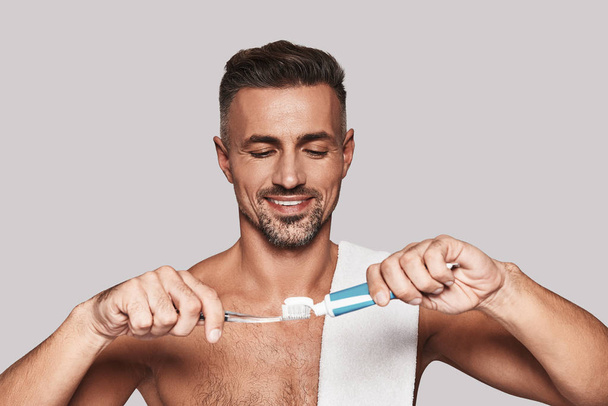 Taking care of his teeth, Handsome young man applying toothbrush and toothpaste while standing against grey background - Foto, Imagem