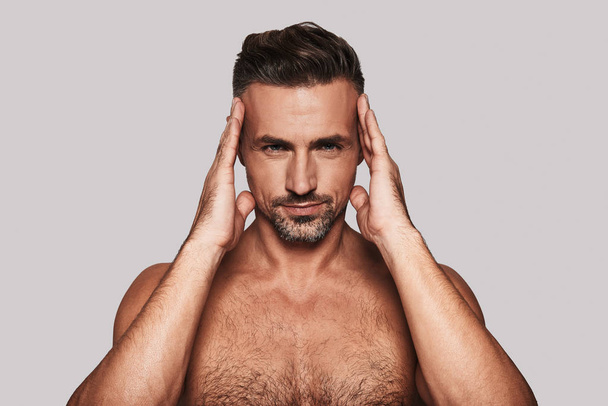 Charming shirtless man keeping hands in hair and smiling while standing against grey background - Foto, immagini
