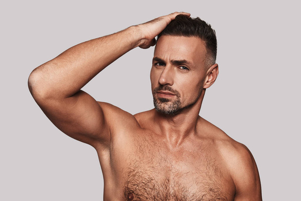shirtless handsome man keeping hand in hair and smiling while standing against grey background - Фото, изображение