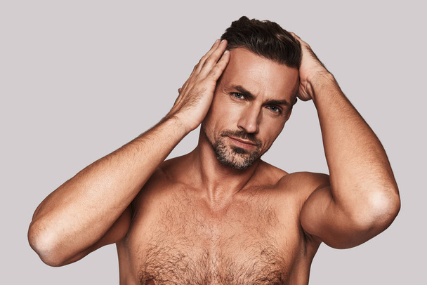Charming shirtless man keeping hands in hair and smiling while standing against grey background - Foto, Imagem