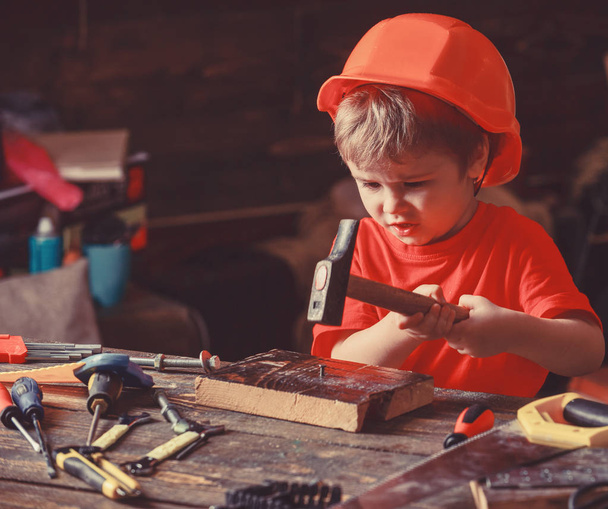 Child in helmet cute playing as builder or repairer, repairing or handcrafting. Toddler on busy face plays at home in workshop. Handcrafting concept. Kid boy hammering nail into wooden board - Foto, afbeelding