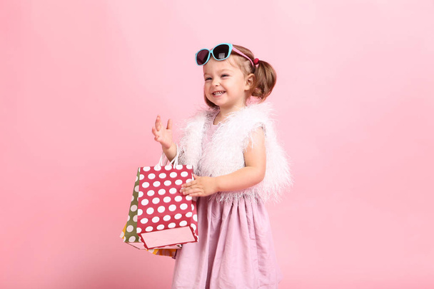 Beautiful little girl in fashion clothes with shopping bags on pink background - Foto, Imagen