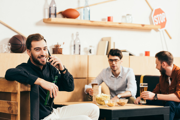 вибірковий фокус усміхненого чоловіка з пивом розмовляє на смартфоні з друзями позаду в кафе
 - Фото, зображення