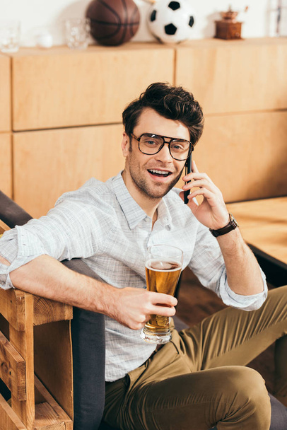 portrait of cheerful man with glass of beer in hand talking on smartphone in cafe - Foto, Imagem