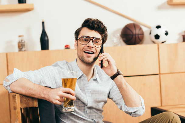 portrait of cheerful man with glass of beer in hand talking on smartphone in cafe - Foto, immagini
