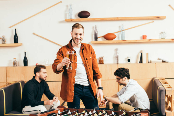 Selektywny fokus z uśmiechnięty człowiek ze szklanką piwa stojący w piłce nożnej tabletu w kawiarni z przyjaciółmi za - Zdjęcie, obraz