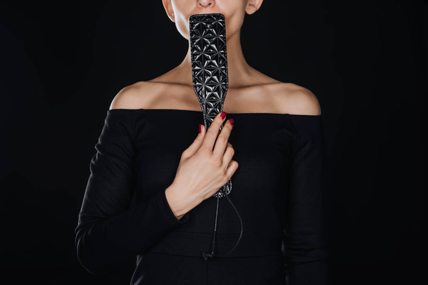gedeeltelijke weergave van vrouw met lederen spanking peddel in de buurt van lippen geïsoleerd op zwart - Foto, afbeelding