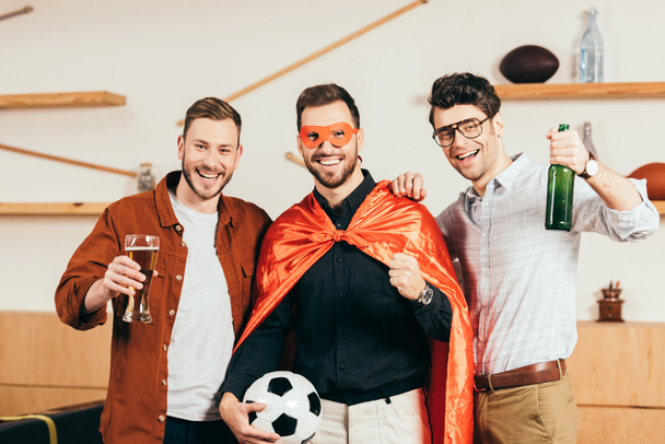 Portret van glimlachende man in superheld kostuum met voetbal in de hand en vrienden in de buurt van door in café - Foto, afbeelding