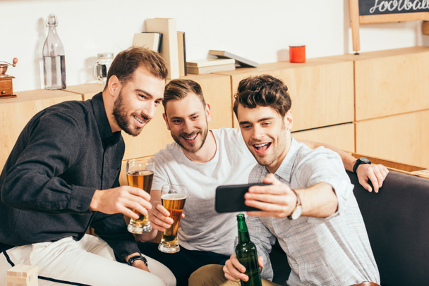 portrait of smiling friends with beer taking selfie on smartphone together in cafe - Fotó, kép