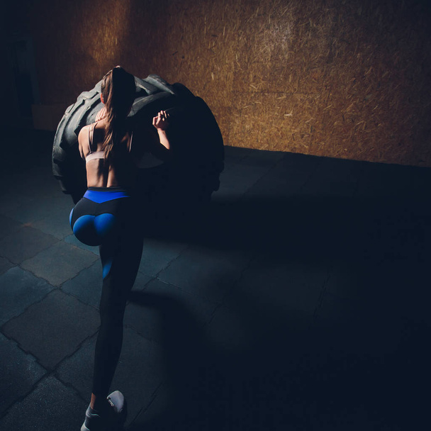 Fitness woman flipping wheel tire in gym. Fit female athlete working out with a huge tire. Back view. Sportswoman doing an strength exercise training. - Photo, Image