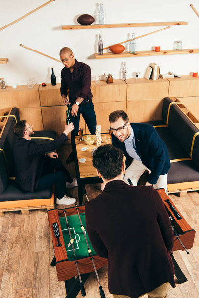 високий кут зору бізнесменів, які грають у настільний футбол, в той час як багатоетнічні колеги смердять напої позаду в кафе
 - Фото, зображення