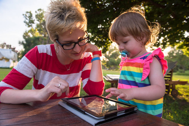 Happy mother and her little daughter enjoying free time using tablet computer while relaxing  on holiday home garden during sunny day - 写真・画像