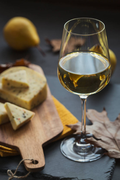 A glass of white wine was served with cheese in a cutting board on dark background. Autumn picnic with cheese, wine and dry leaves in rustic style. - Photo, image