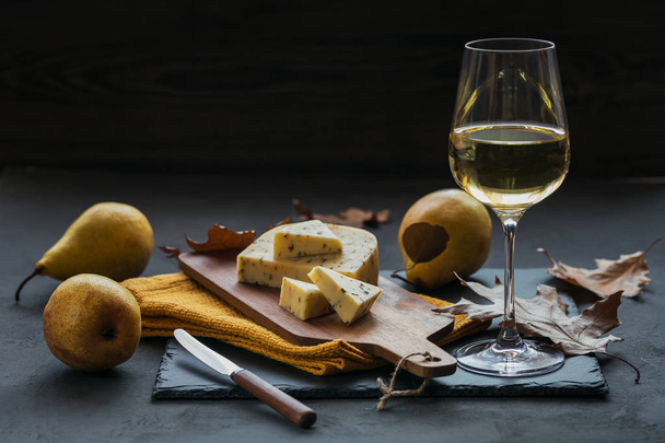 A glass of white wine was served with cheese in a cutting board on dark background. Autumn picnic with cheese, wine and dry leaves in rustic style. - Photo, Image