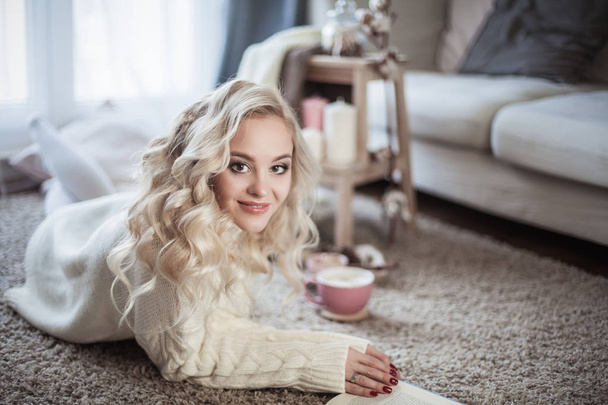 Cozy home portrait of a young beautiful woman. A beautiful blonde girl in a knitted dress is sitting by the window drinking a cappuccino and reading a book. Autumn decor. Cozy. Winter. Autumn. - Foto, immagini