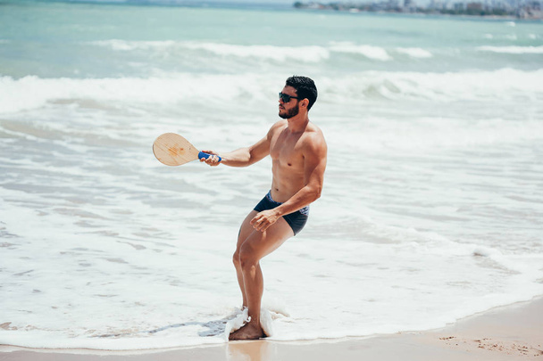 Young Brazilian man playing beach tennis on the beach with swimwear - Photo, image