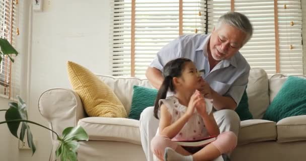 Asian senior grandfather tying little girl hair while she reading book at home. - Filmagem, Vídeo