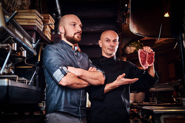 Kuchař, čerstvý steak a jeho asistent, stojící vedle v kuchyni restaurace. - Fotografie, Obrázek