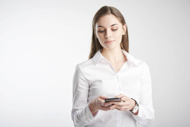 Beautiful young businesswoman wearing white shirt and text messaging while standing against at isolated white background. - Fotografie, Obrázek