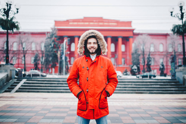 Portret, makro młody stylowo człowiek uśmiechający się z brodą ubrany w Czerwona zimowa kurtka z kapturem i futro na jego głowę stoi na tle miasta pokryte śniegiem. Kompozycja zima i mróz - Zdjęcie, obraz