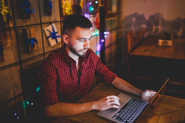 Portrait of a young handsome Caucasian man with a beard and toothy smile in a red checkered shirt sitting beside a laptop at a wooden table. In the evening at the coffee shop - Photo, Image