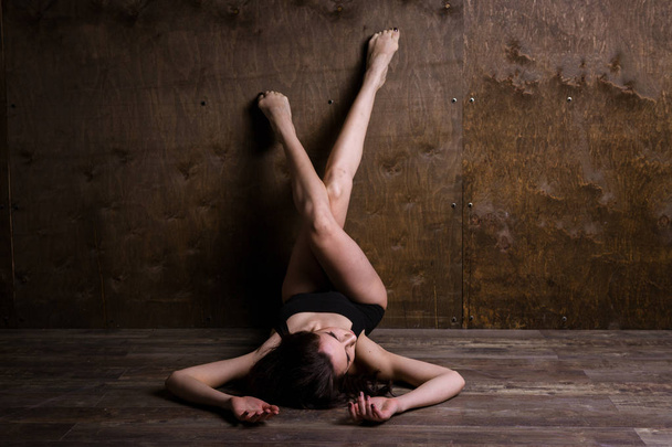 Theme is a sport and a beautiful sexy woman. Young caucasian girl lying on the floor kicking up posing in a black sports swimsuit clothes with bare feet on a dark wooden background in the studio. - Fotografie, Obrázek