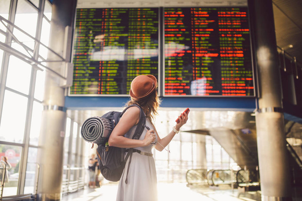 Motyw podróży i tranosport. Piękna młoda kaukaski kobieta w sukni i plecak stojąc wewnątrz dworca kolejowego lub terminalu, patrząc na harmonogram, trzymając czerwony telefon, używa technologii komunikacyjnych. - Zdjęcie, obraz