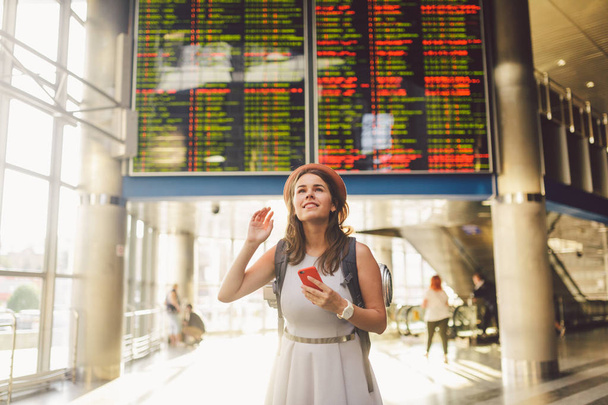 Utazási téma és tranosport. Gyönyörű fiatal kaukázusi nő a ruhát, és állt befelé vasútállomás, illetve a terminál látszó-on egy menetrend gazdaság egy piros telefon, hátizsák, használja a kommunikációs technológia. - Fotó, kép