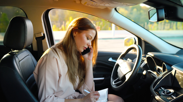 Mujer de negocios hablando en un teléfono inteligente y tomando notas en el diario sentado en el coche
 - Metraje, vídeo