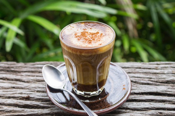 Latte káva ve skle s lžící na dřevěné tabulce na pozadí přírodní Zelený strom. Latte káva v kavárně nebo na caf - Fotografie, Obrázek