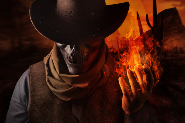 Foto de un vaquero de cabeza de cráneo demoníaco con sombrero sosteniendo un hechizo de fuego con su brazo sobre un fondo desierto al atardecer
. - Foto, imagen