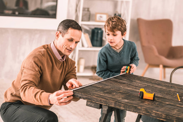 Занятой серьезный человек просит своего малолетнего сына помочь измерить стол
. - Фото, изображение