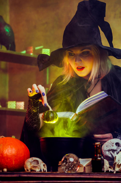 Image of witch blonde in black hat with book brewing potion in pot with green steam - Foto, Imagem