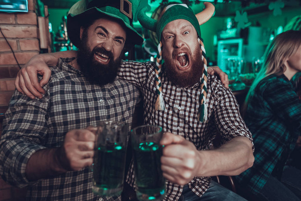Saint Patrick's Day Party. Friends is Celebrating. People is Drinks a Green Beer. Friends is Young Men and Woman. Beard Men Screaming and Toast with Each Other. Men Wearing a Green Hats. Pub Interior. - 写真・画像