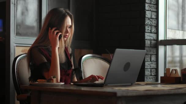 Młoda kobieta siedzi w kawiarni w drewniany stół, picia kawy i za pomocą smartfonu. Na stole jest laptop. Dziewczyna, przeglądania Internetu, rozmowy, blogów. Kobieta trzyma telefon i patrząc na jego ekran. - Materiał filmowy, wideo