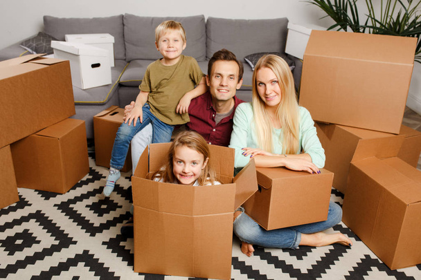 Foto de pareja feliz con hijo e hija sentados en el suelo entre cajas de cartón
 - Foto, Imagen
