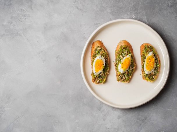 Top view of three whole-grain toasts plated avocado with eggs, fresh mini herbs, sunflower seeds and sprinkled black salt served on white ceramic plate - Foto, Imagem
