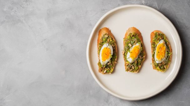 Top view of three whole-grain toasts plated avocado with eggs, fresh mini herbs, sunflower seeds and sprinkled black salt served on white ceramic plate - Foto, imagen