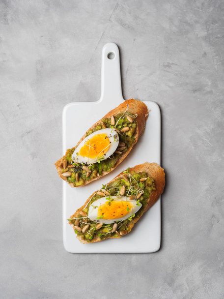 Top view of whole-grain toasts plated avocado with eggs, fresh mini herbs, sunflower seeds and sprinkled black salt served on ceramic white board - Zdjęcie, obraz